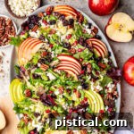 top down look at apple salad made with brussels sprout, apples, cranberries and topped with a vinaigrette. oval serving bowl is sitting on a gray surface with cut apples around it.