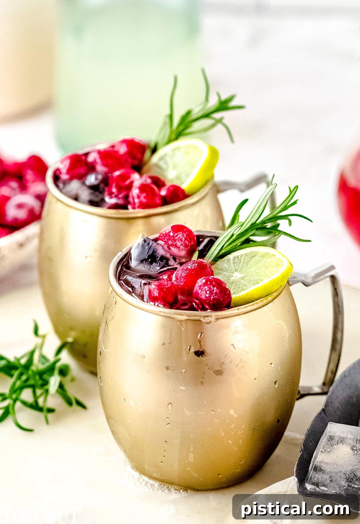 Two Cranberry Moscow Mule cocktails on a white board, garnished with fresh cranberries, lime slices, and a sprig of rosemary, exuding holiday cheer.