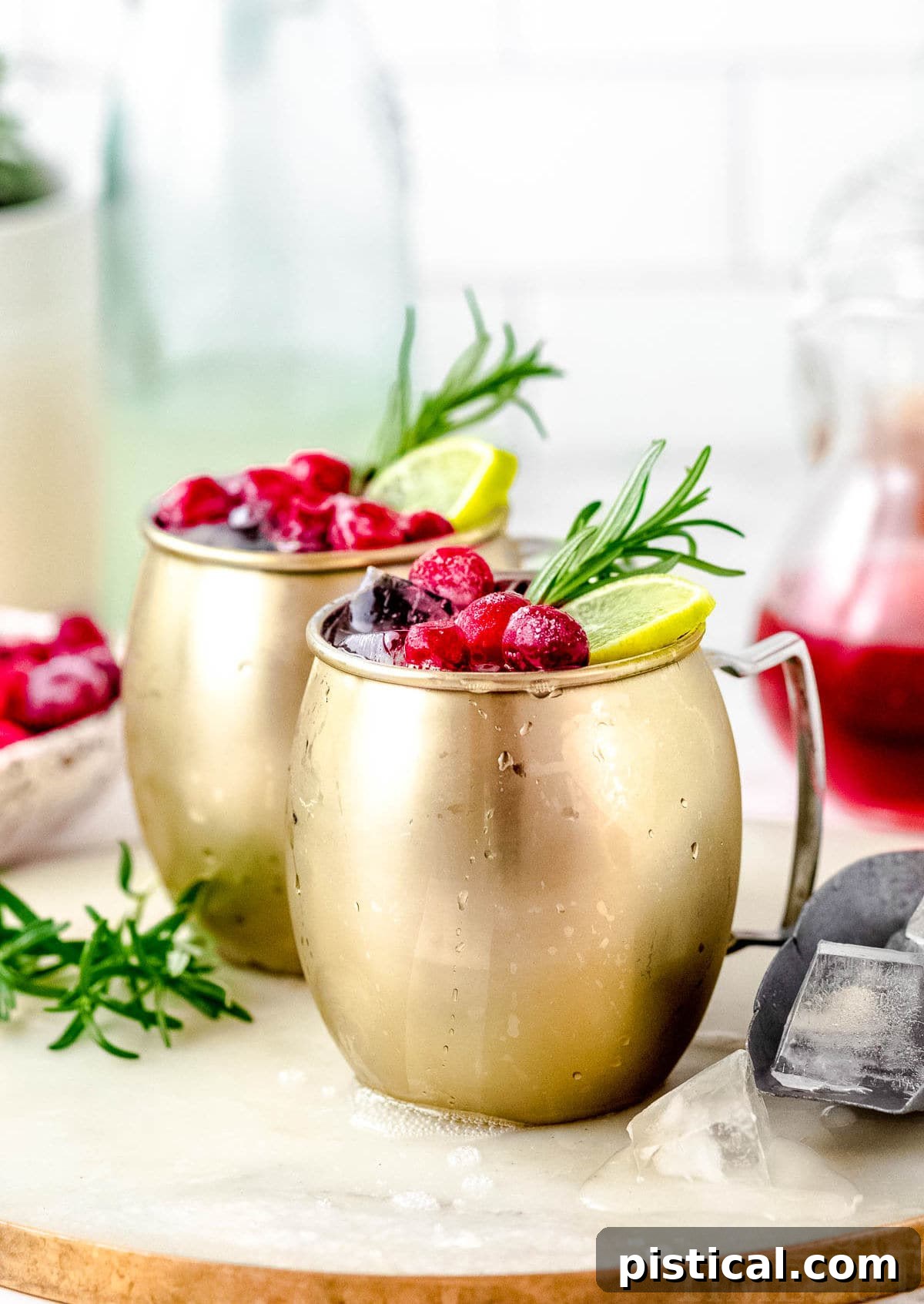 Two vibrant Cranberry Moscow Mule cocktails arranged on a clean white surface, beautifully garnished with fresh cranberries, lime slices, and fragrant rosemary sprigs.