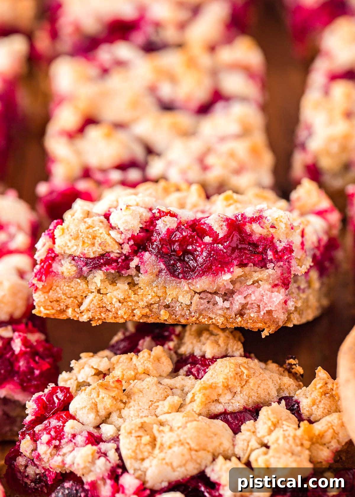 Close-up of golden brown Cranberry Bars on a wooden board, with one bar lifted to showcase the vibrant red cranberry filling and crumbly topping.