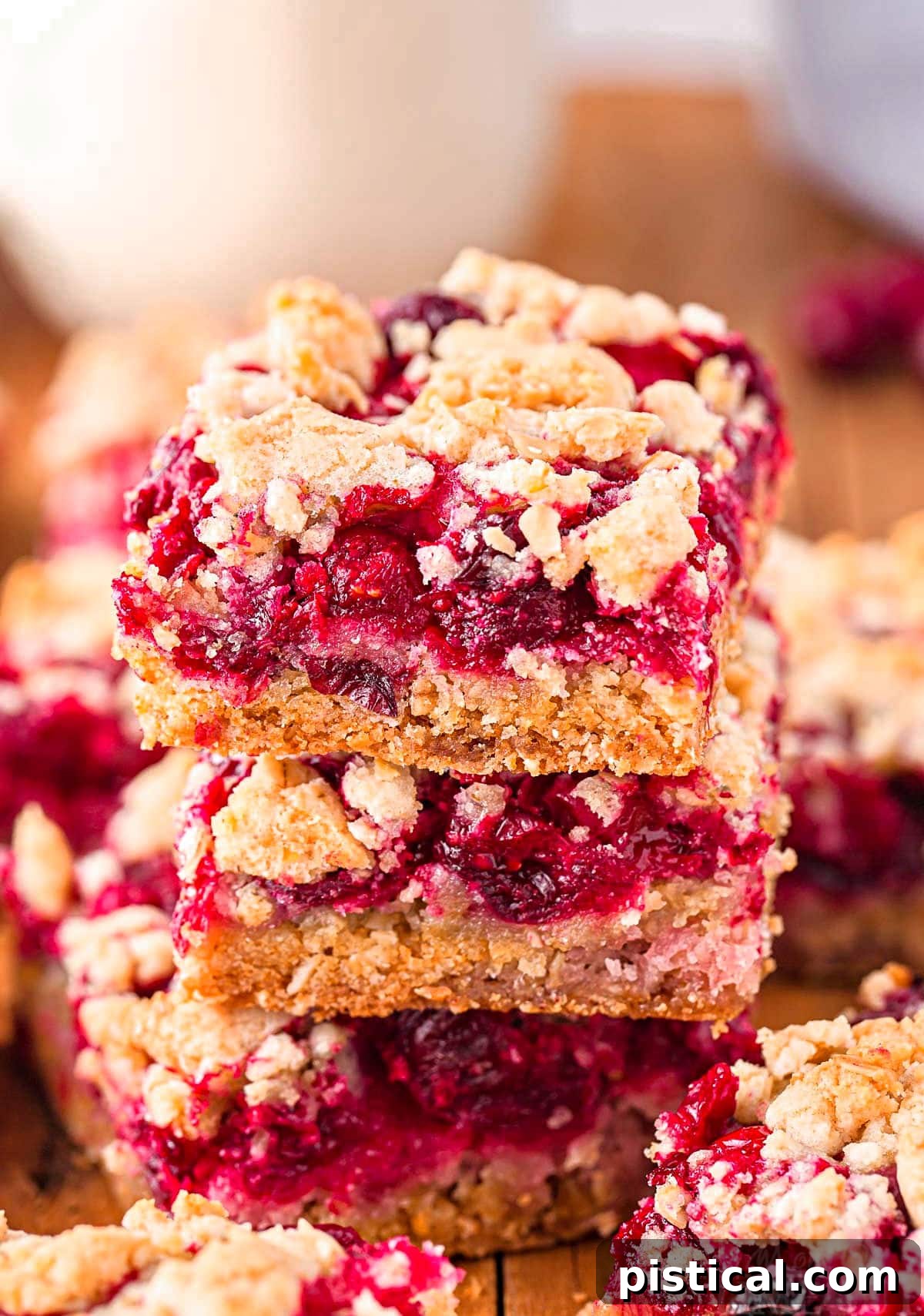 A stack of three homemade Cranberry Crumb Bars, showcasing the layers of buttery crust, sweet-tart filling, and golden crumble, placed on a dark wooden board with a glass of milk in the background.