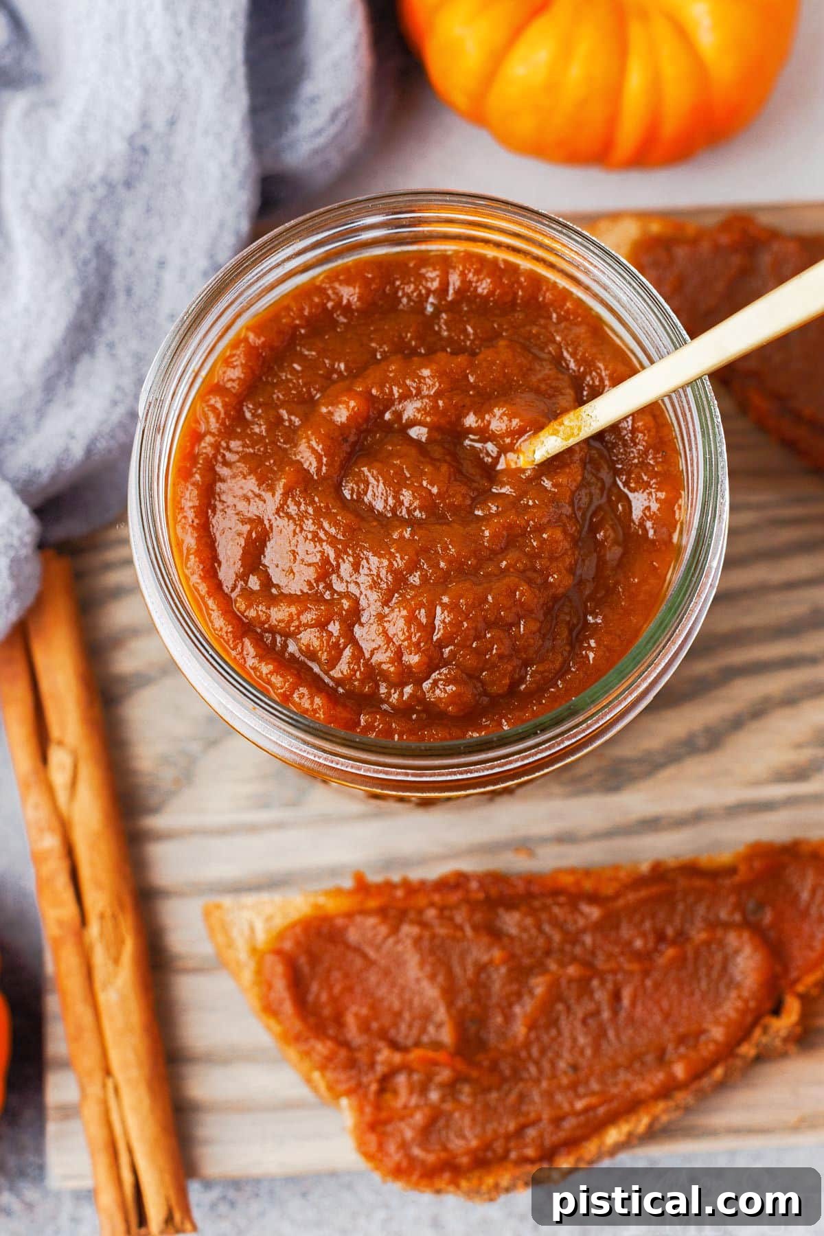 top down view of jar of pumpkin butter with small gold spoon sitting on a gray board with a half piece of toast with pumpkin butter slathered on it.