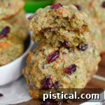 Zucchini cookies on a wood board with white rim. Top cookie has been halved. Carrot and zucchini can be seen in the background.