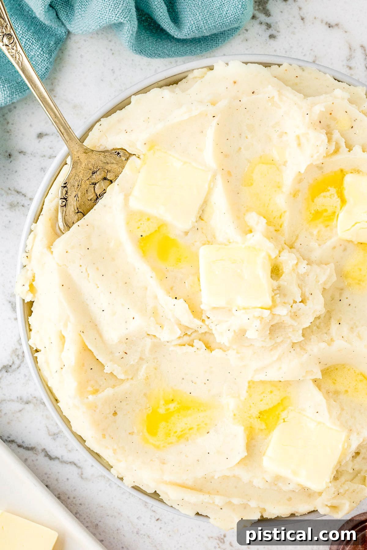 A shallow bowl of perfectly creamy Instant Pot Mashed Potatoes, generously topped with a pat of melting butter and a sprinkle of freshly ground black pepper. A teal kitchen towel is visible in the background, adding a touch of color to the inviting scene.