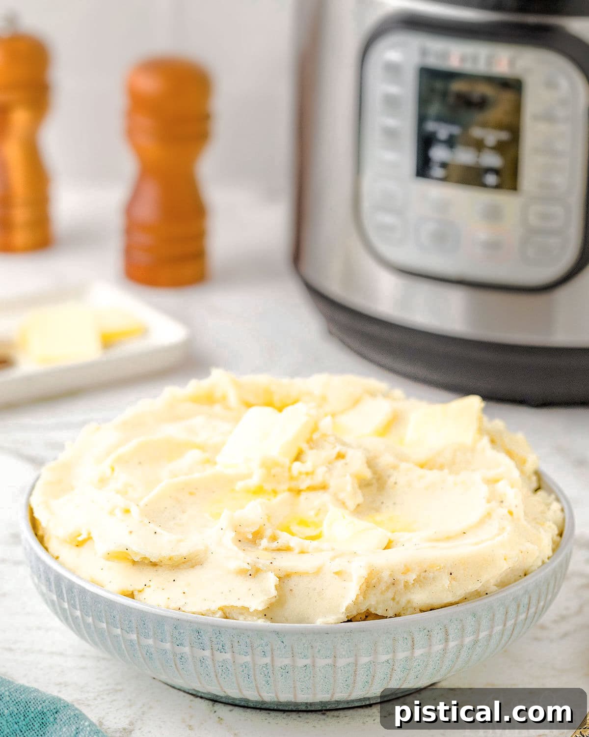 A cozy scene featuring a bowl of perfectly mashed potatoes resting in front of a modern Instant Pot. Salt and pepper grinders sit nearby, ready for seasoning.