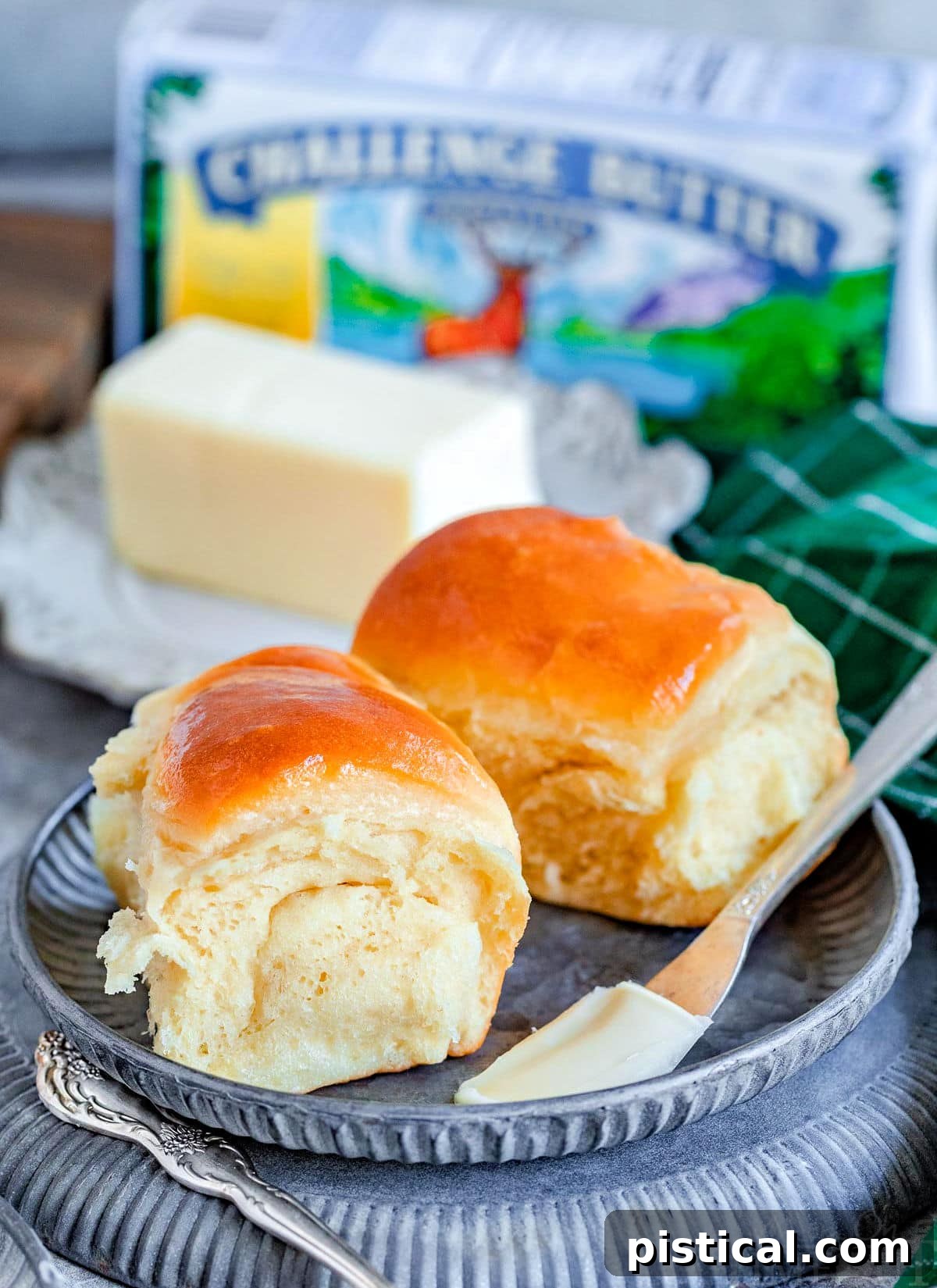 two parker house dinner rolls on a metal plate with a stick of butter behind them. a box of butter can be seen in the background.