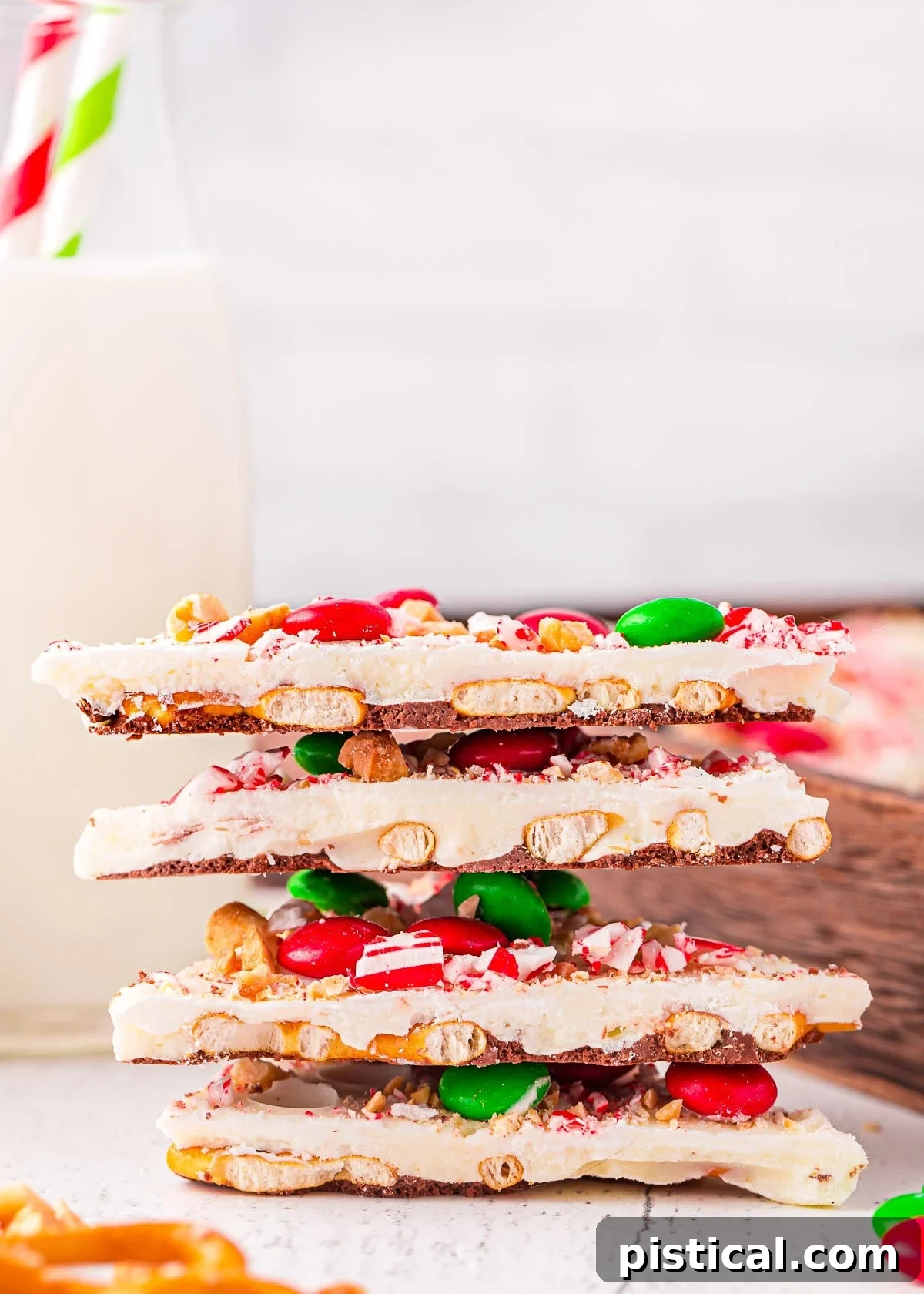 four pieces of christmas bark stacked on top of each other. the bark is made with pretzels, m and ms, and candy canes. glass of milk in background.