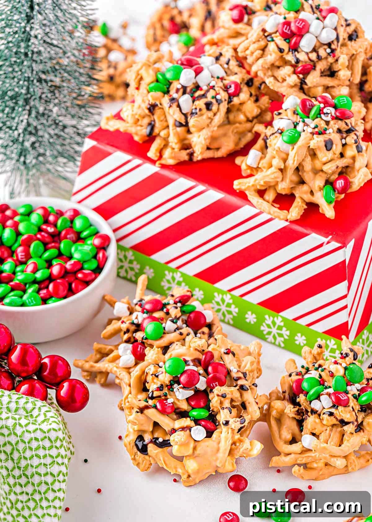 Butterscotch haystacks, beautifully decorated for Christmas, arranged on and around a red and white striped Christmas box, with a small bowl of red and green M&M candies beside them.