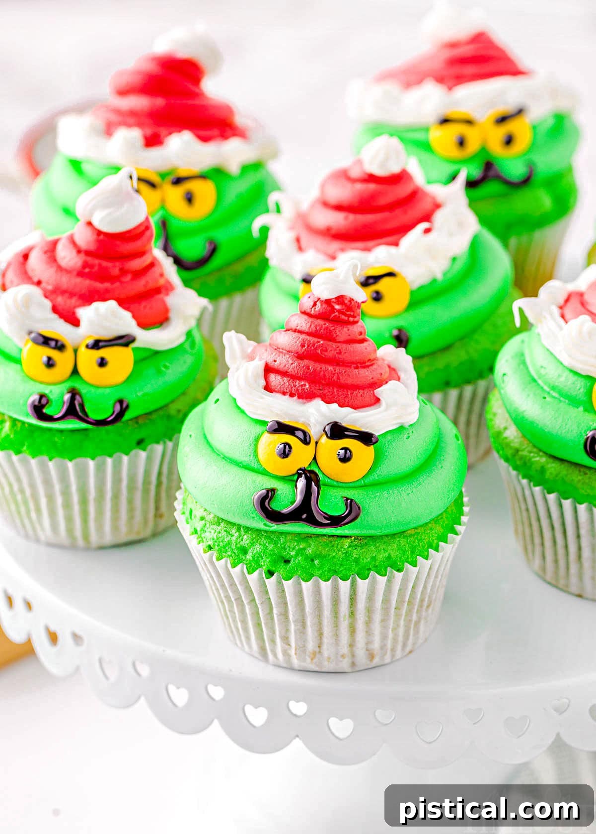 Six vibrant green Grinch cupcakes, each topped with a red Santa hat and a cheerful Grinch face, arranged artfully on a white cake stand. These festive Christmas treats are perfect for holiday parties.