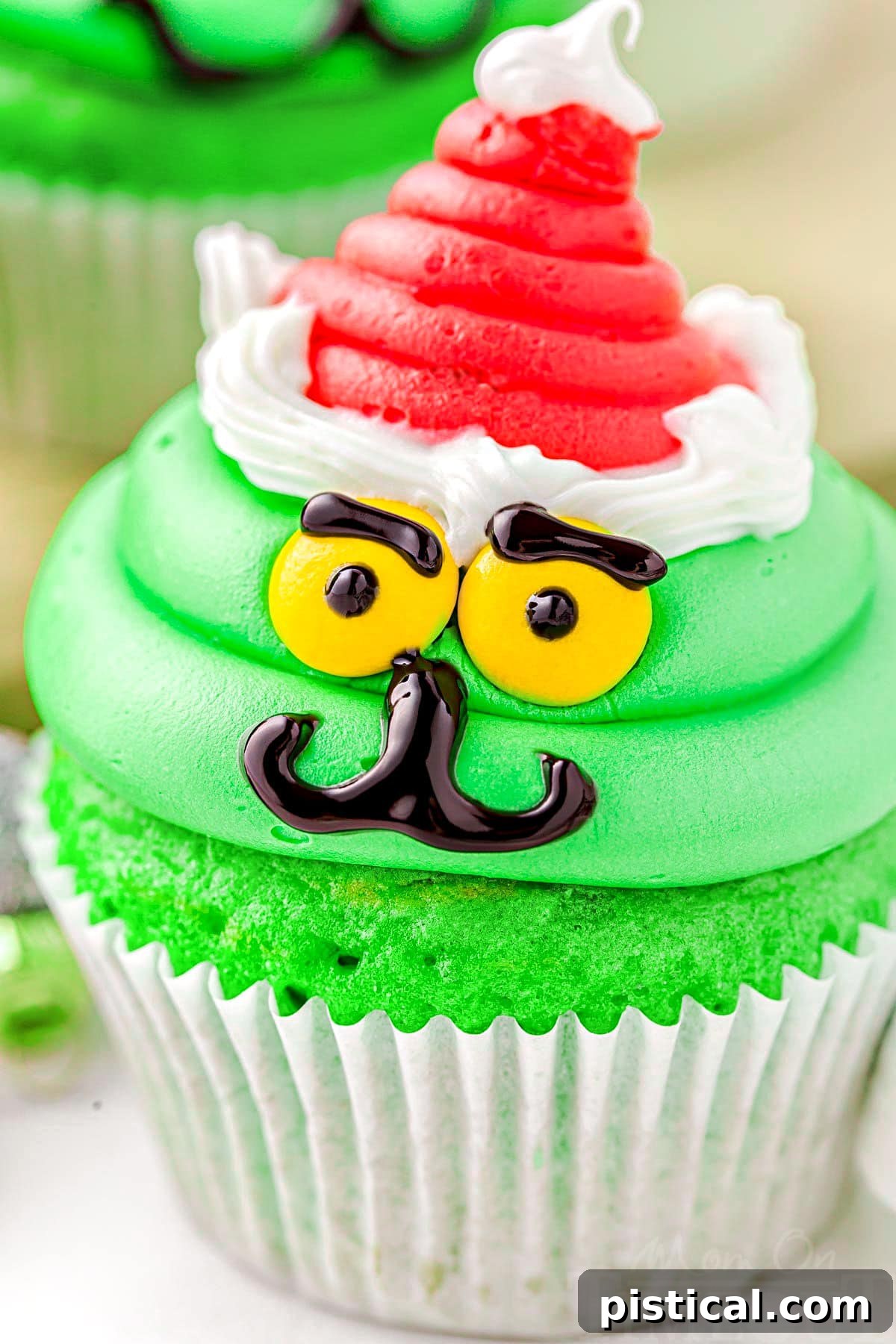 A close-up view of a single Grinch cupcake, showcasing its bright green frosting, detailed facial features, and a perfectly piped red Santa hat with white trim, sitting on a pristine white background.