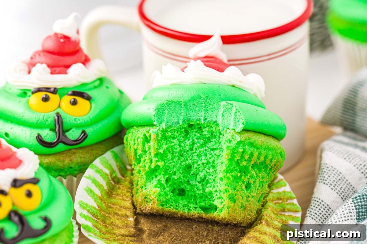 A selection of green cupcakes frosted and decorated like the Grinch, with one cupcake featuring a bite taken out of it, set against a festive holiday backdrop.