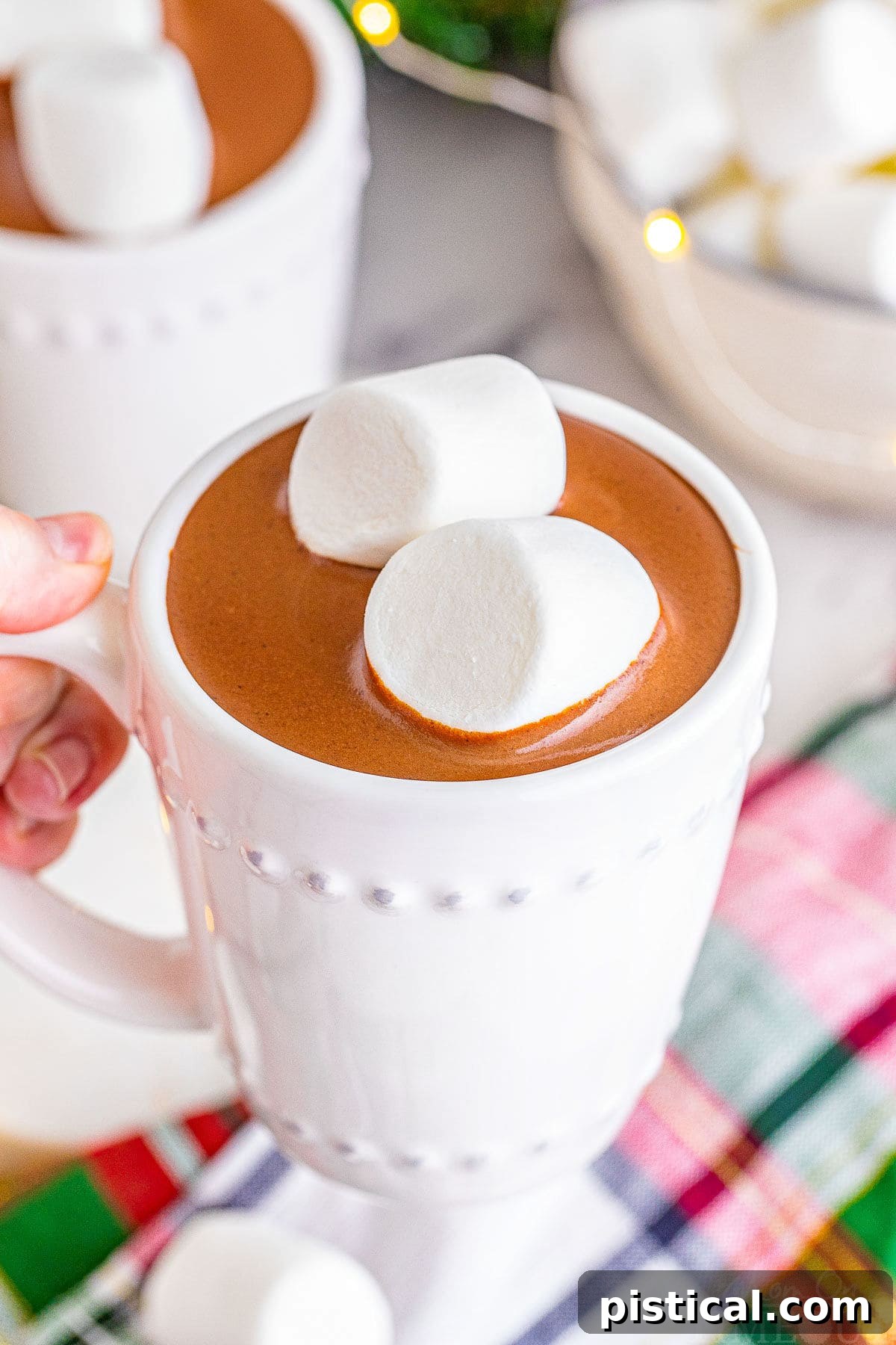 A white mug filled with decadent crockpot hot chocolate, topped with two jumbo marshmallows, held over a festive Christmas linen.
