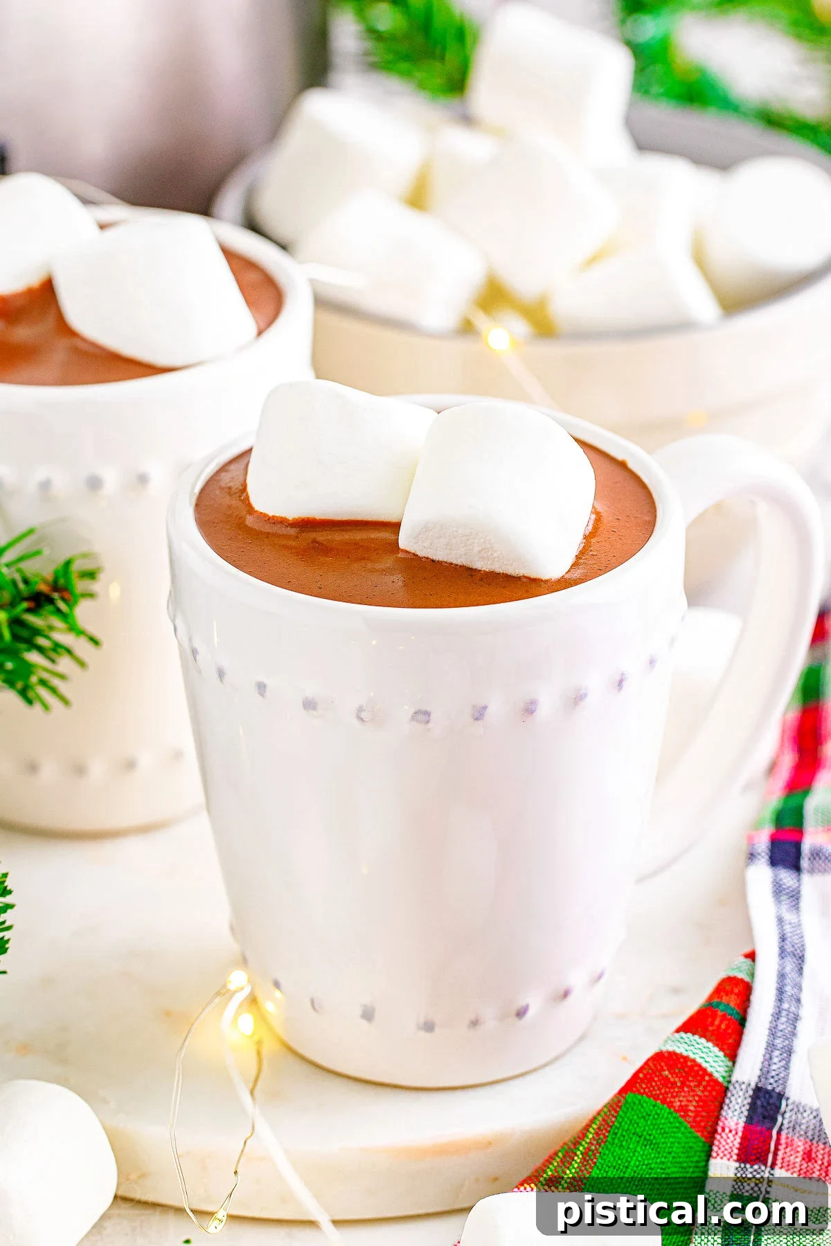 A white mug filled with hot chocolate and topped with marshmallows, positioned in front of another mug of crockpot hot cocoa.