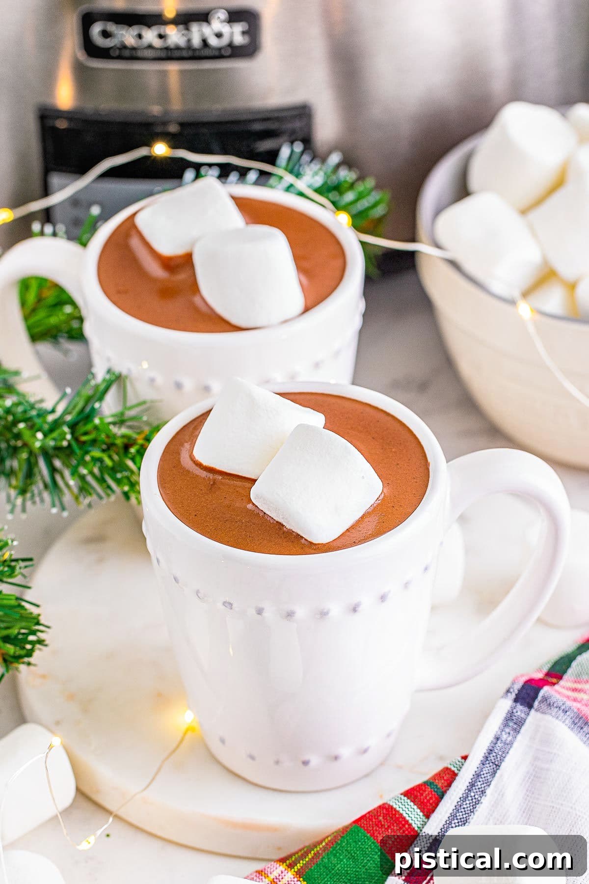 Two cups of hot chocolate, topped with marshmallows, sitting in front of a silver crockpot.