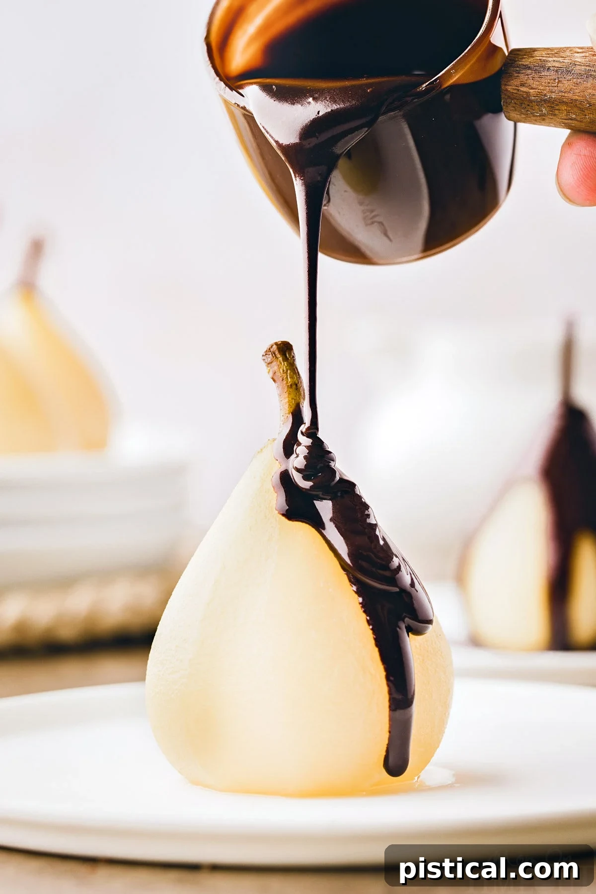 Velvet Pear Indulgence 2 poached pear sitting on white plate with chocolate sauce about to being drizzled onto the pear.