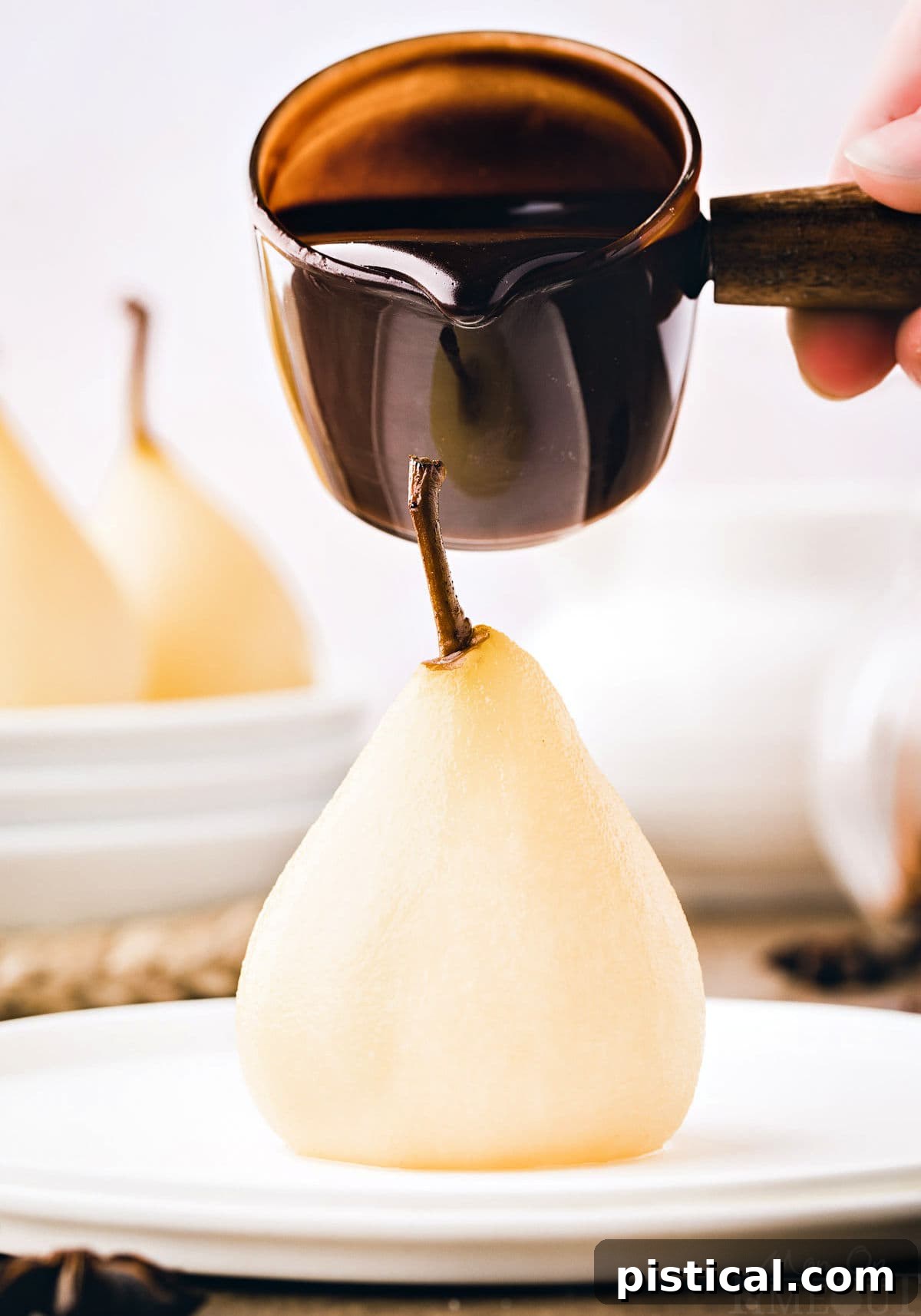 Velvet Pear Indulgence 7 poached pear sitting on white plate with chocolate sauce about to being drizzled onto the pear.