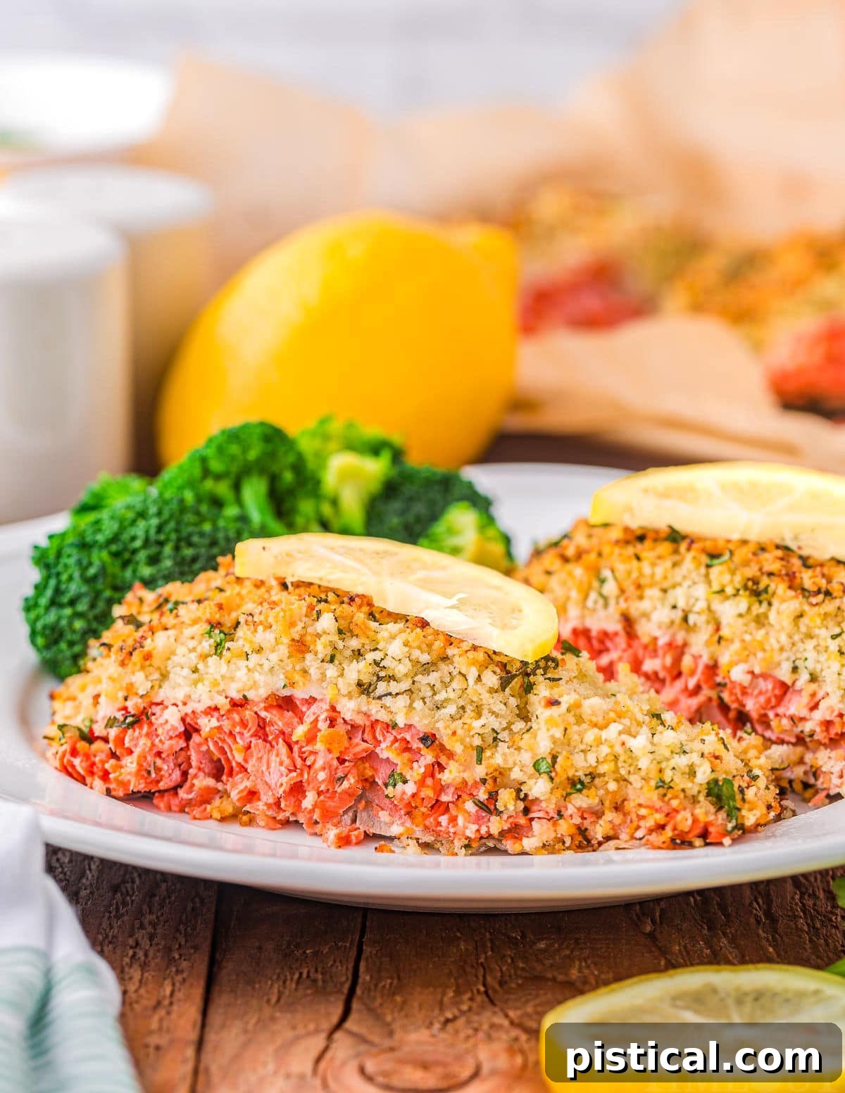 Crispy Parmesan Herb Salmon Bake 2 Two pieces of oven-baked salmon with a crispy Parmesan herb crust, garnished with fresh lemon slices, on a white plate. Lemon slices are also visible in the background and foreground.