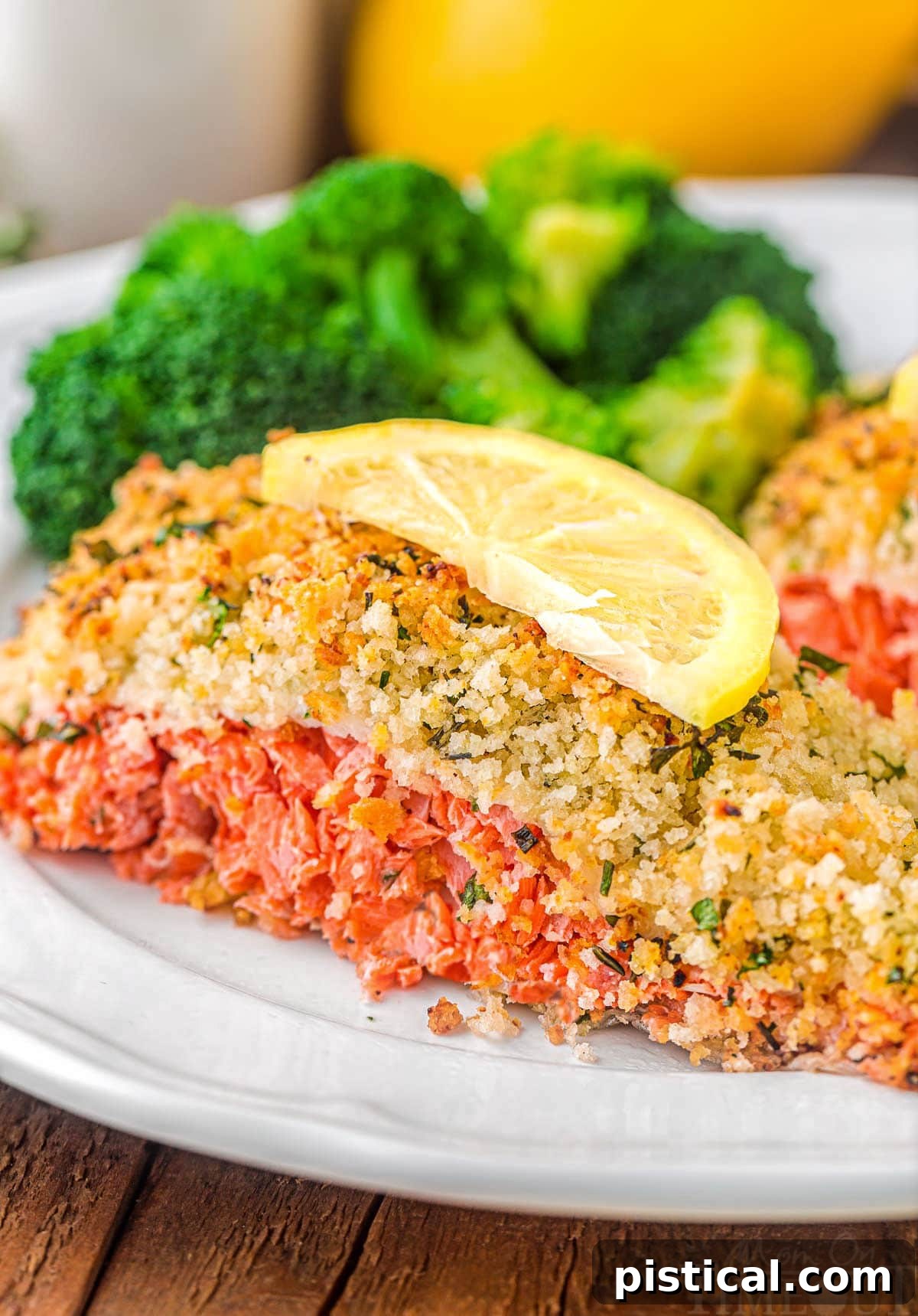 Crispy Parmesan Herb Salmon Bake 7 A vibrant piece of baked salmon, adorned with a crunchy Panko crust, served on a white plate with fresh broccoli and a bright lemon slice resting on top.