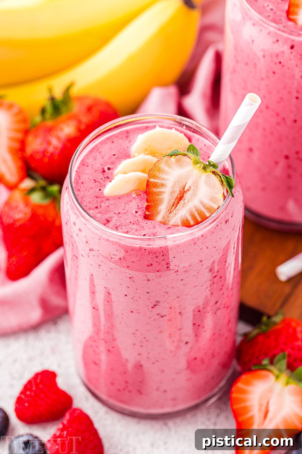 A refreshing strawberry banana smoothie in a tall glass, garnished with a half strawberry and banana slices. A pink striped straw is ready for sipping, with fresh strawberries and bananas blurred in the background, suggesting natural goodness.