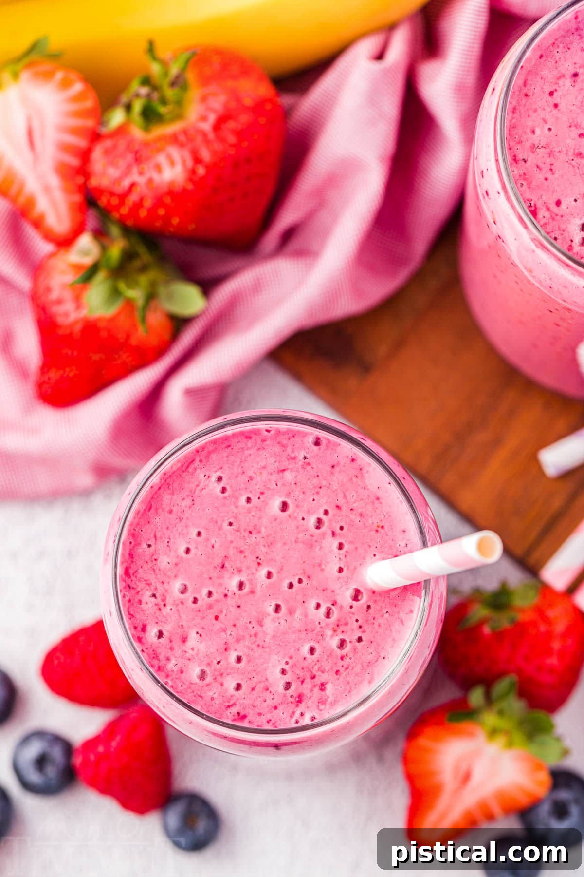 A top-down view of a vibrant strawberry banana smoothie served in a clear glass cup with a charming pink and white striped paper straw. More fresh strawberries and banana slices are artfully scattered around the base of the glass, completing the inviting presentation.