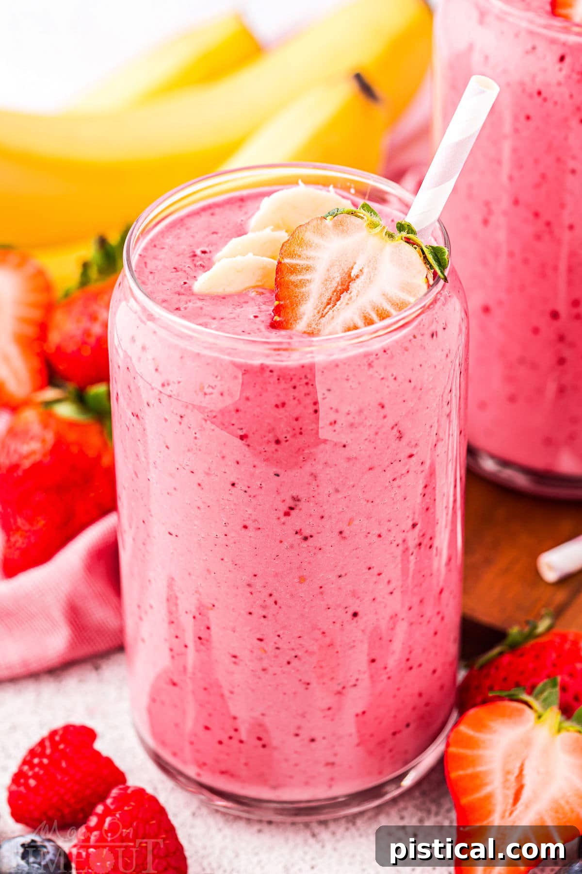 A vibrant strawberry banana smoothie in a tall glass, complete with a charming pink and white striped straw. The background features a soft blur of fresh bananas and strawberries, highlighting the natural, delicious ingredients.