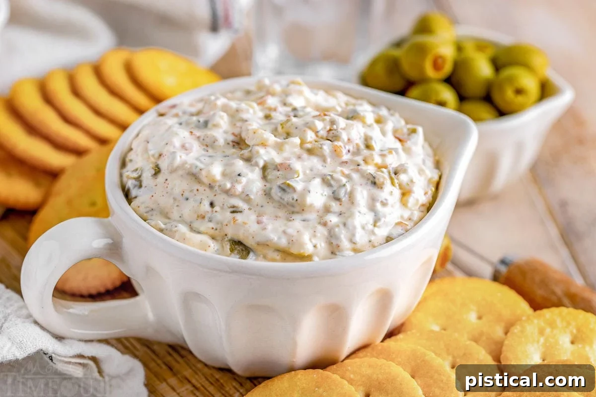 Creamy green olive dip served in a white bowl, elegantly arranged on a wooden board with various crackers. A small bowl of whole green olives sits in the soft background.