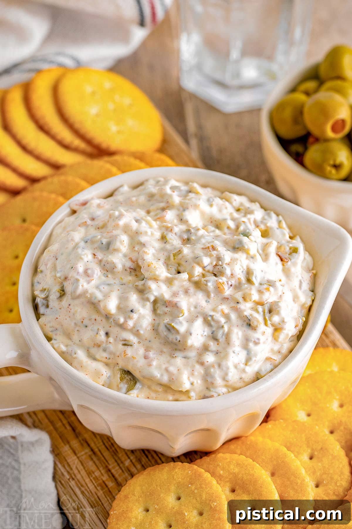 A top-down angled view of a white bowl filled with creamy green olive dip, garnished with olive slices. Ritz crackers are neatly arranged on a wooden board beside the dip, ready for serving.