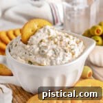 Creamy olive dip in a small white bowl sitting on a wood board surrounded by crackers and a small bowl of green olives sitting off to the side. A glass of water in the background.