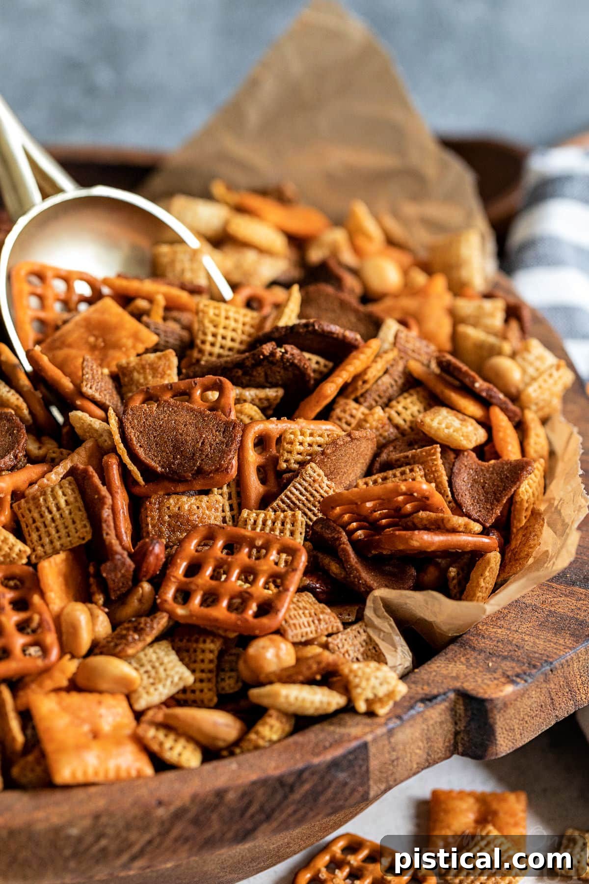 A large wooden bowl, lined with brown parchment paper, overflowing with golden-brown homemade Chex Mix. A large gold scoop is playfully sticking out of the mix, inviting you to grab a handful.