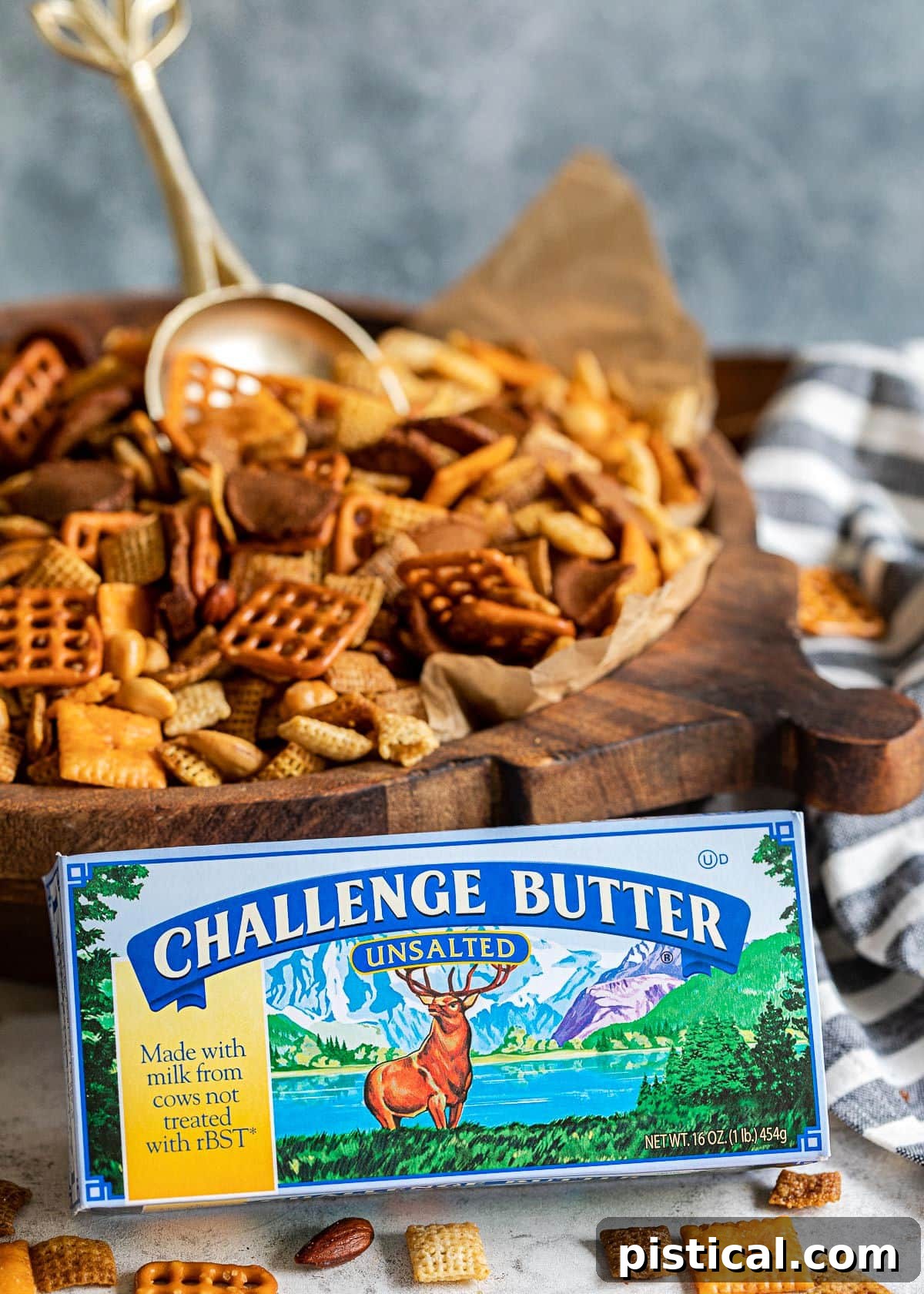 A box of Challenge butter sits prominently in the foreground, with a large wooden bowl of homemade Chex Mix and a gold scoop visible in the soft-focus background.