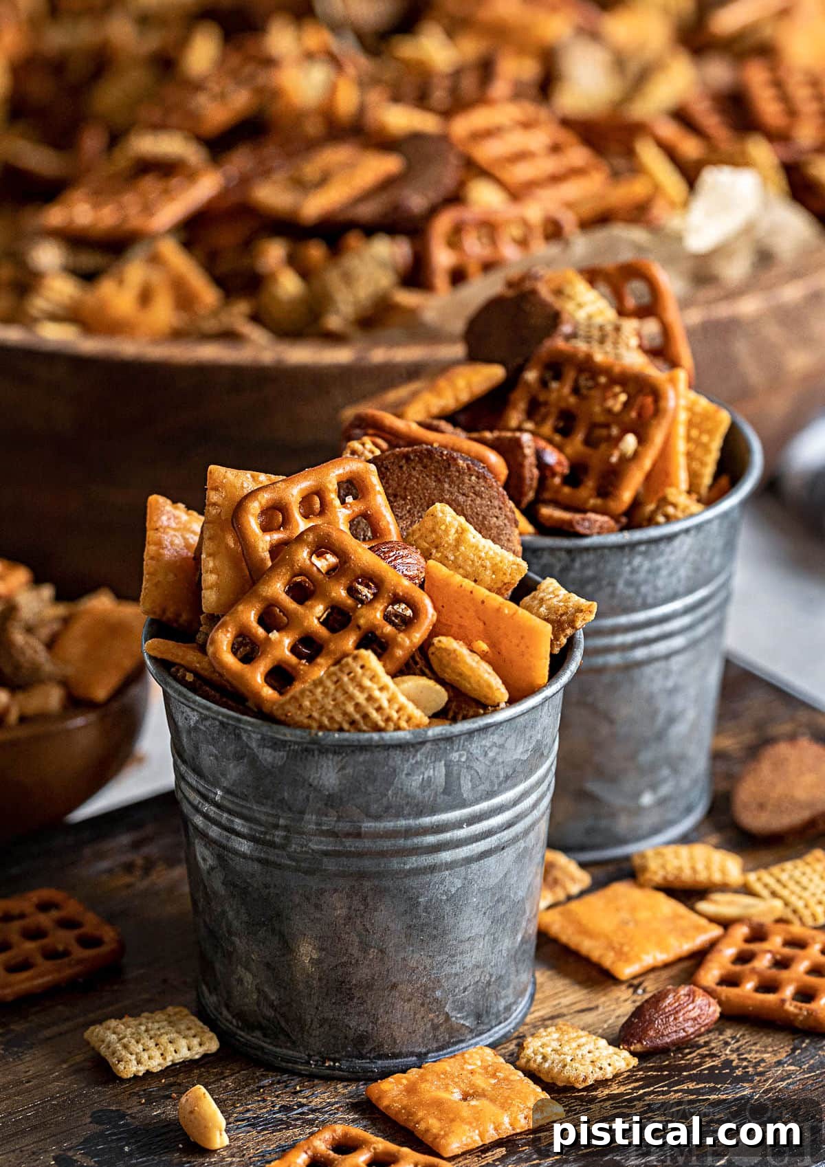Homemade Chex Mix recipe artfully arranged in small metal serving containers, with a larger wooden bowl brimming with the remaining mix in the blurred background, creating an inviting scene.