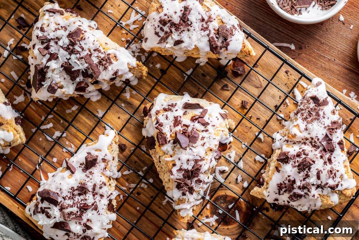 A top-down view of five exquisitely iced Chocolate Coconut Scones, adorned with dark chocolate shavings, resting on a cooling rack atop a wooden board, showcasing their perfect golden hue.