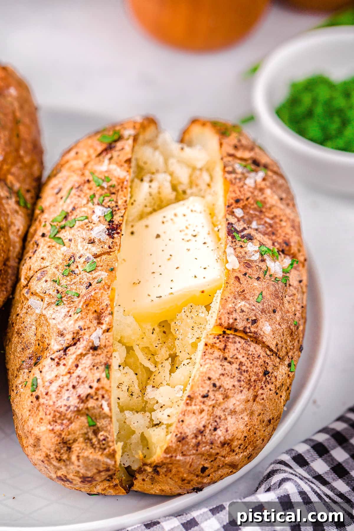 Perfect Air Fryer Baked Potatoes 2 Air fryer baked potato on a white plate topped with butter, salt and pepper. Freshly chopped green chives are in a small white bowl in the background.