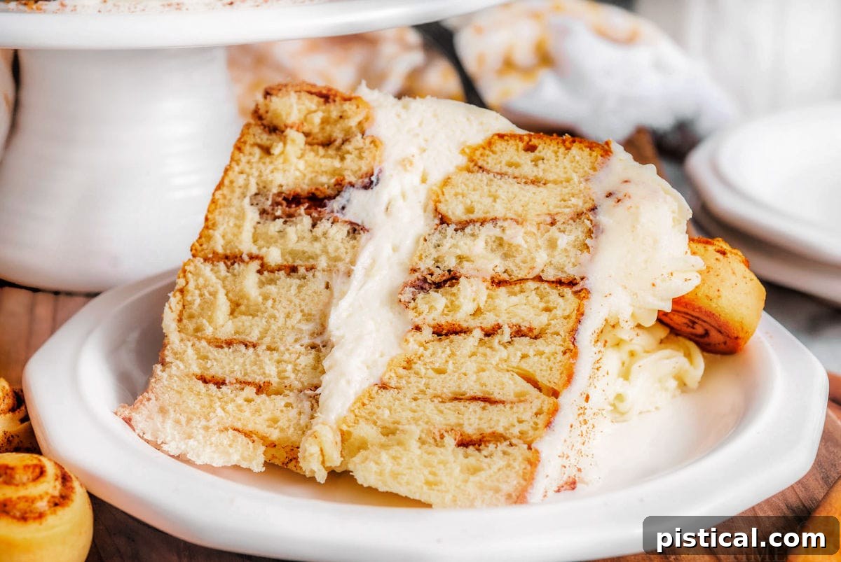 Decadent Cinnamon Swirl Layer Cake 5 A generous slice of Cinnamon Roll Layer Cake rests on a white plate, with the rest of the cake displayed on a white cake stand in the softened background, highlighting its inviting appearance.