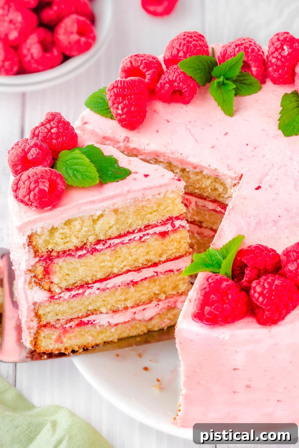 A beautiful slice being carefully removed from a four-layer vanilla cake, revealing luscious raspberry buttercream and vibrant raspberry filling between each moist layer. The cake is elegantly adorned with fresh raspberries and delicate mint leaves.