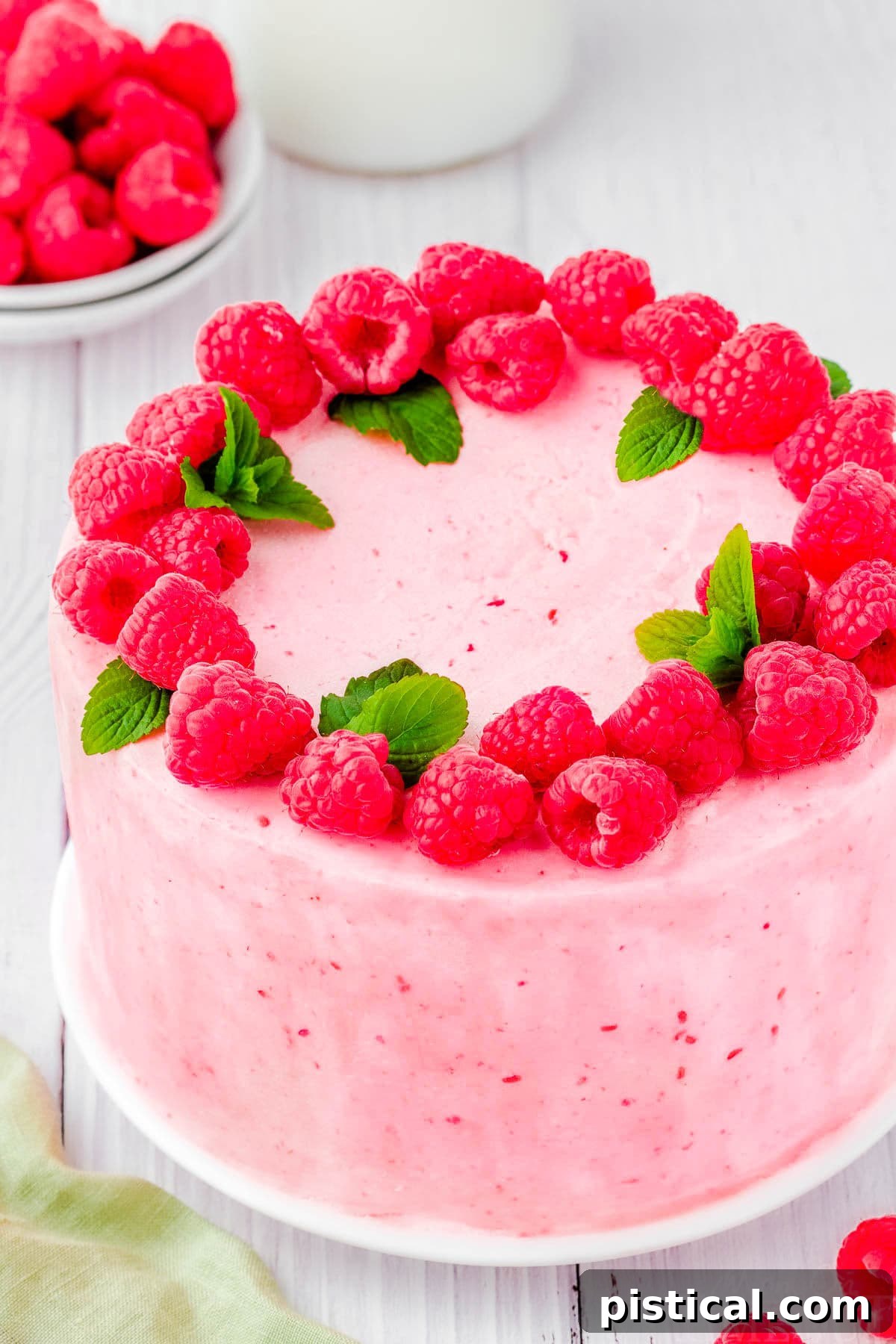 A magnificent whole vanilla cake adorned with generous swirls of raspberry buttercream and a lavish topping of dozens of fresh raspberries and sprigs of mint, ready to be served.