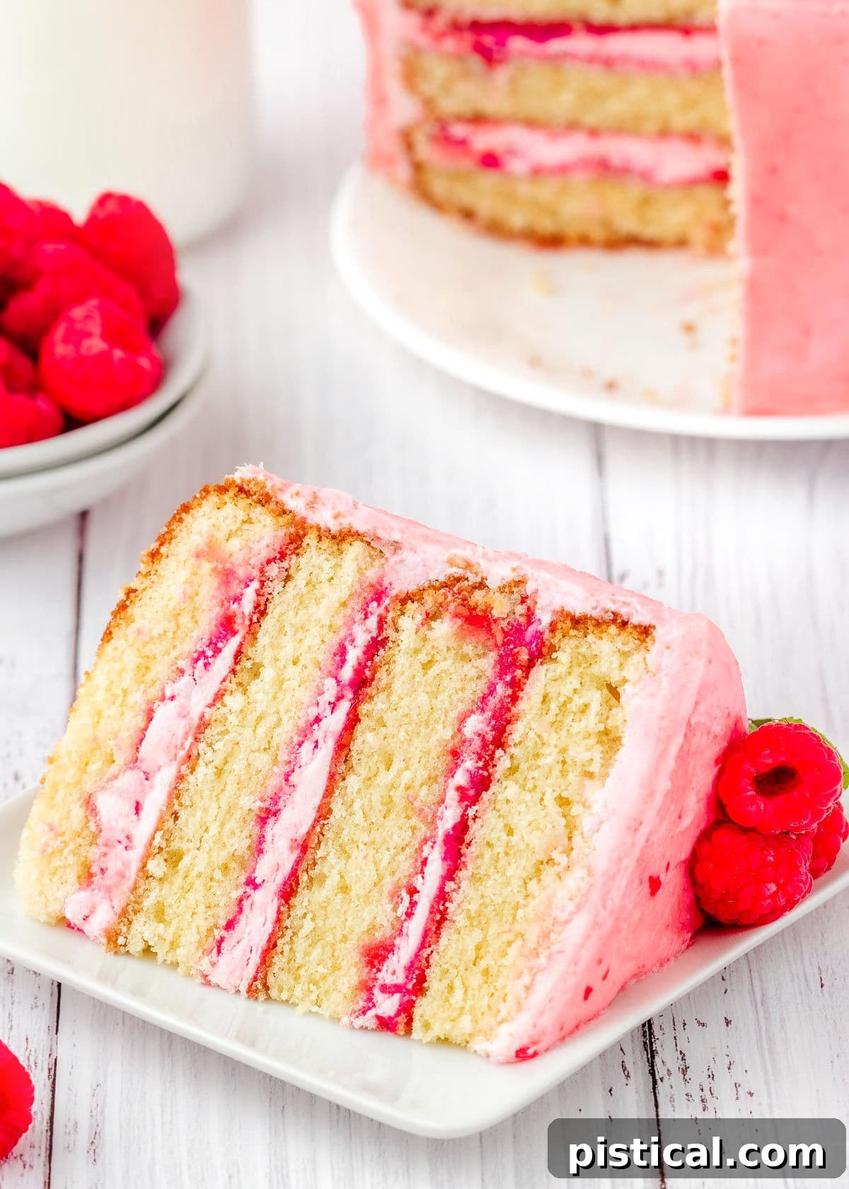 A perfectly portioned slice of Raspberry Vanilla Cake gracefully displayed on a crisp white square plate, positioned invitingly in front of the remaining tiered cake.