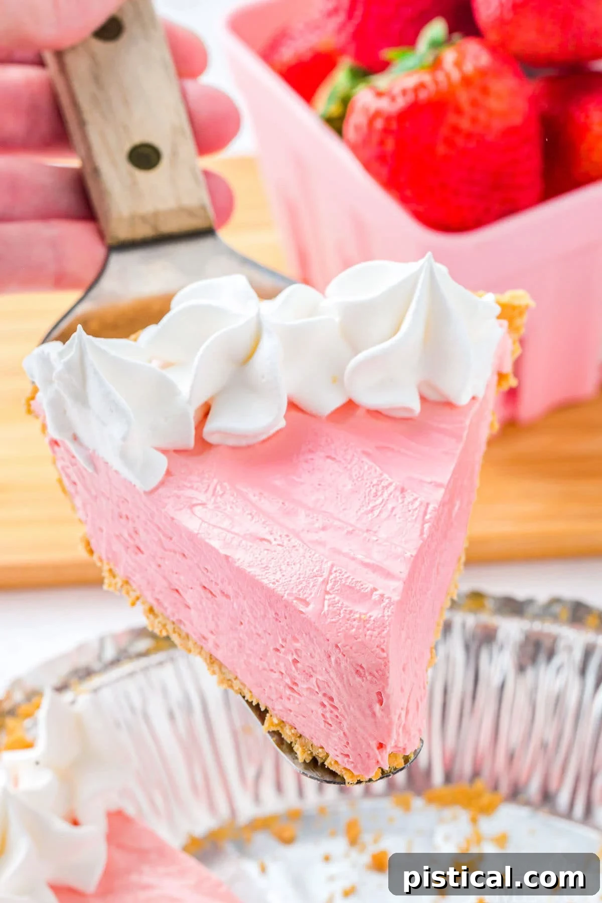 A vibrant slice of strawberry Kool Aid Pie, generously topped with fluffy whipped cream and a fresh strawberry, held above the full pie.