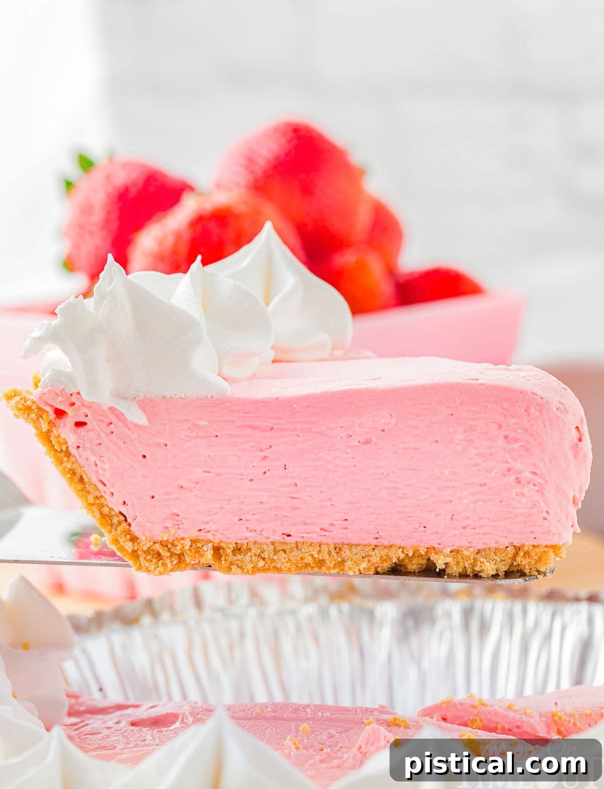 A server holds a perfect pink slice of Kool Aid Pie, topped with fresh strawberries and whipped cream, invitingly above the rest of the beautifully set pie.