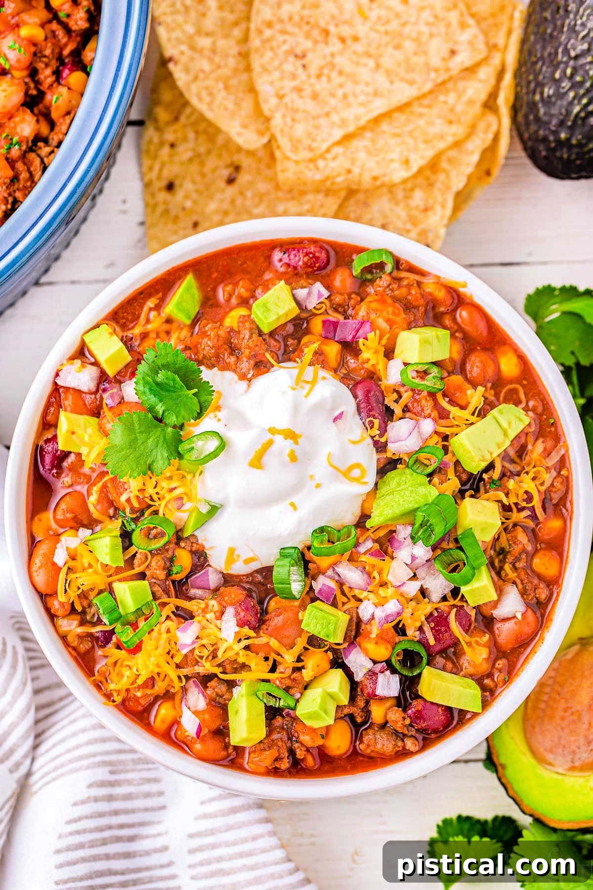 top down look at a generous serving of taco soup in a white bowl topped with avocado, sour cream and cheese. tortilla chips and other toppings can be seen around the bowl.