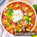 top down look at a generous serving of taco soup in a white bowl topped with avocado, sour cream and cheese. tortilla chips and other toppings can be seen around the bowl.
