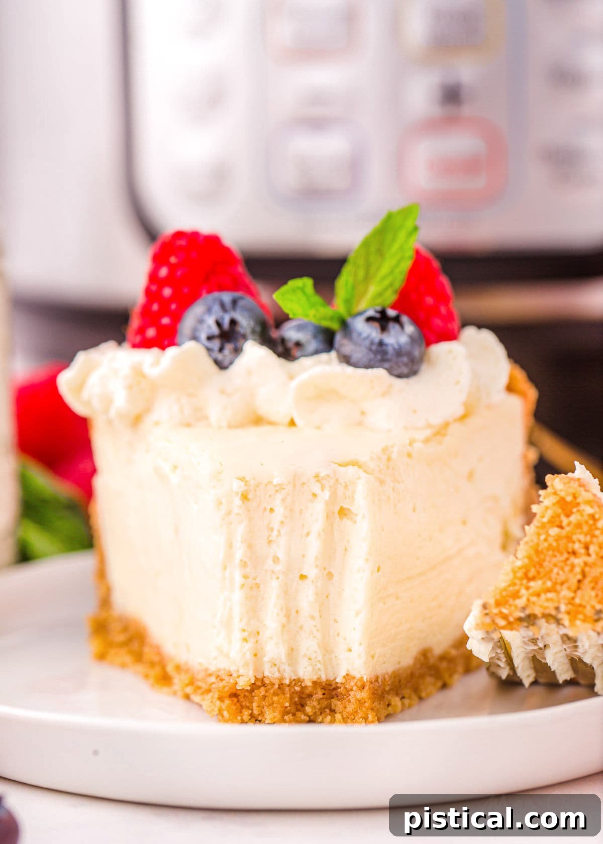 A delectable piece of Instant Pot cheesecake on a small, round white plate, with a forkful already removed. The cheesecake is beautifully topped with fresh berries and a generous dollop of whipped cream, showcasing its irresistible appeal.