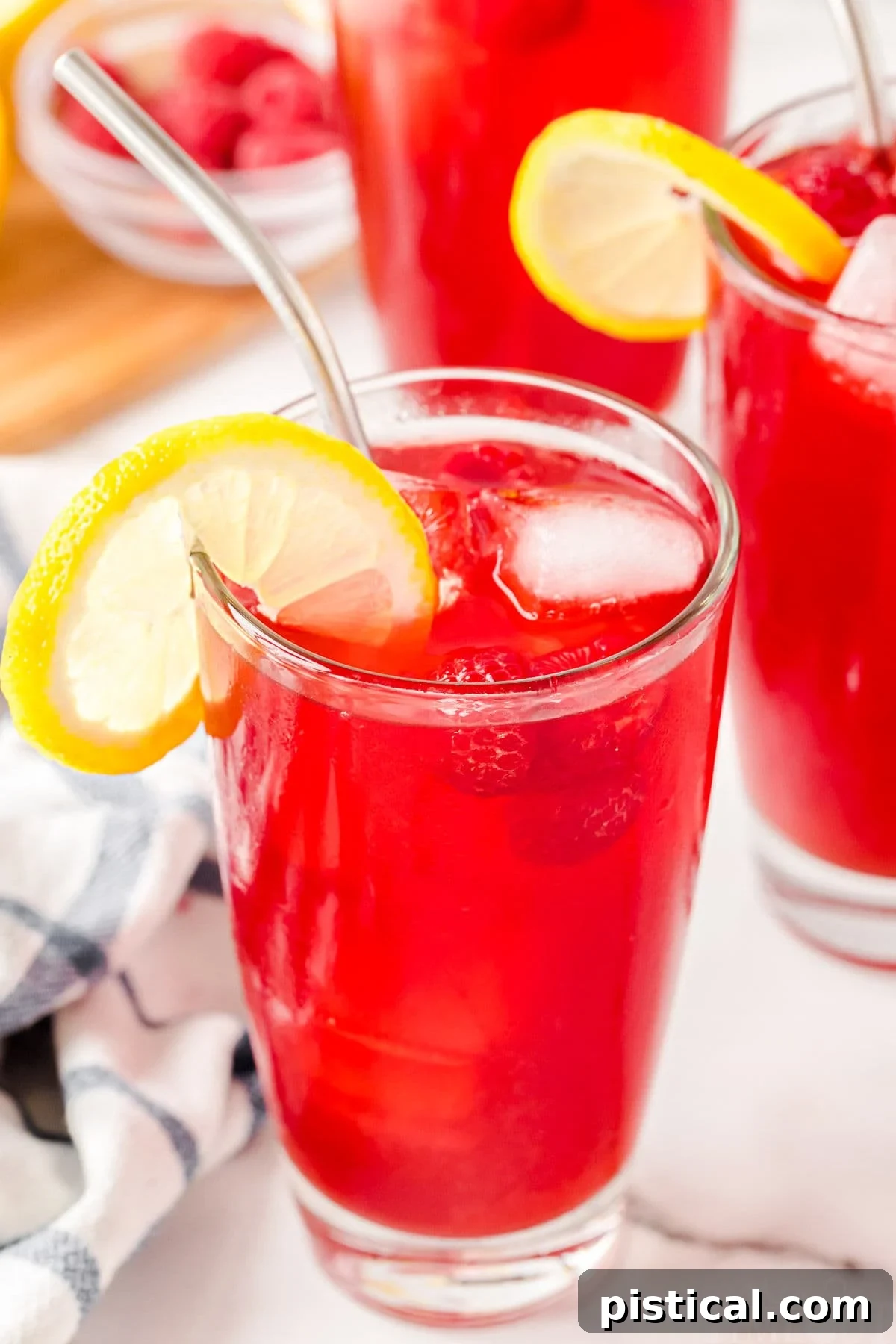 Ruby Red Raspberry Refresher 3 top down angled view of a glass filed with vibrantly colored raspberry iced tea topped with a lemon slice. a stainless steel straw is inserted into the glass and two more filled glasses can be seen in the background.