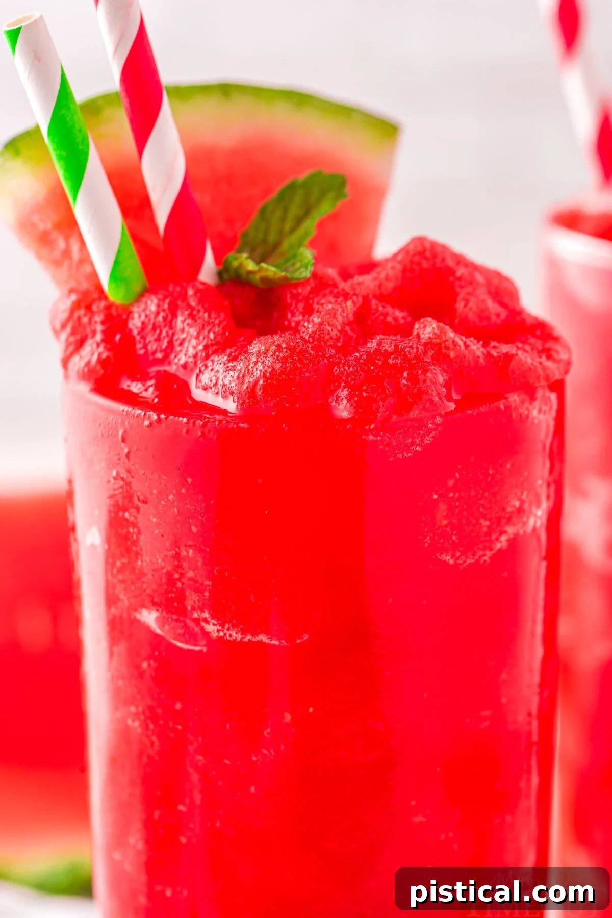 A close-up view of a tall, clear glass filled to the brim with a bright red watermelon slushie, elegantly garnished with a fresh sprig of mint, a watermelon wedge, and two paper straws.