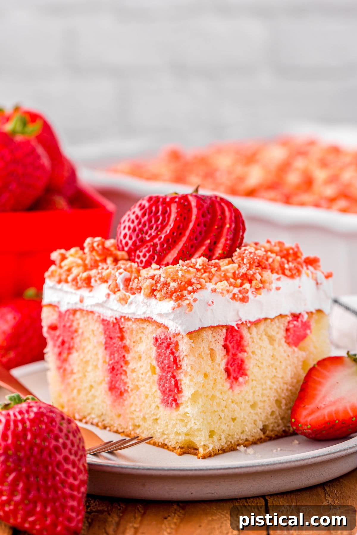 serving of strawberry poke cake on small white round plate topped with fresh sliced strawberry and crunch topping. rest of cake can be seen in background.