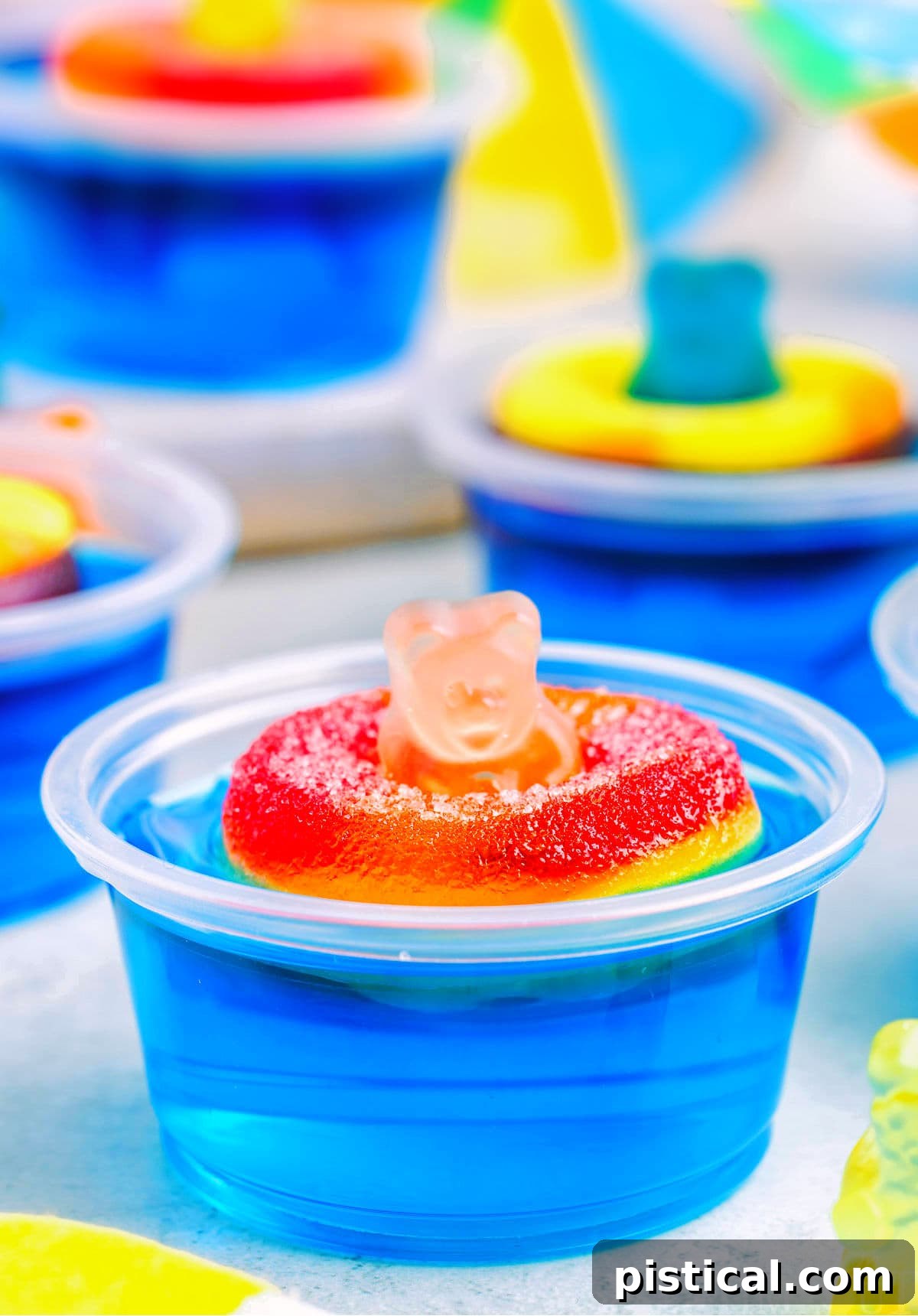 blue pool party jello shot with gummi bear sitting on white surface. more jello shots can be seen in the background.
