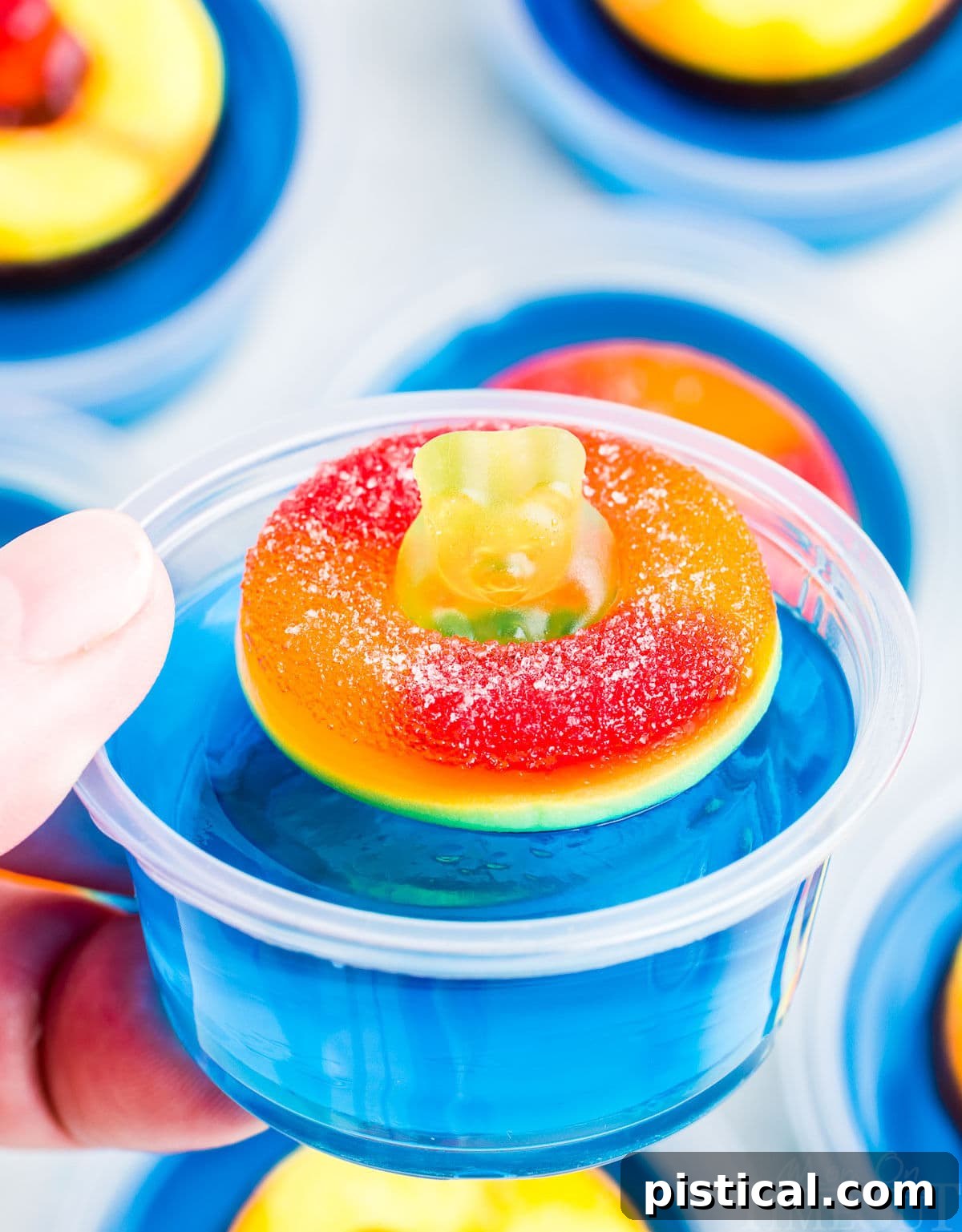 hand holding single blue jello shot with a peach ring and gummy bear on top. more jello shots can be seen in the background.