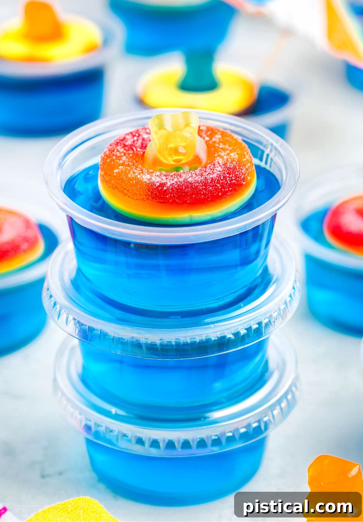 three blue jello shots stacked with lids. top shot has a peach ring and gummy bear on it.