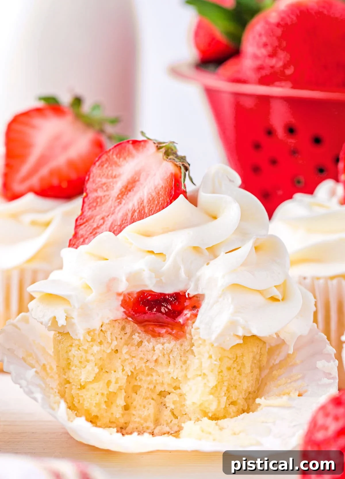 Vanilla cupcake with strawberry jam filling and creamy buttercream frosting, topped with half a fresh strawberry. A bite has been taken, revealing the vibrant strawberry jam inside.