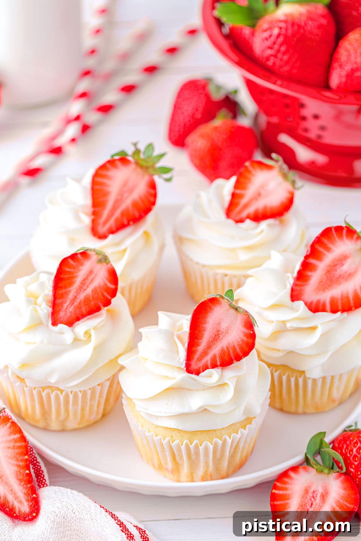 Top-down view of five vanilla cupcakes, each filled with strawberry jam and topped with creamy vanilla buttercream and half a fresh strawberry, arranged on a white plate.